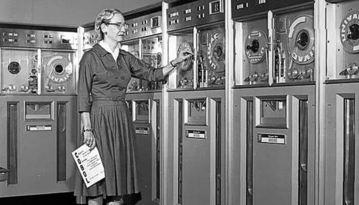 Grace Hopper examinando unidades de fita magnética do UNIVAC com um manual de programação COBOL nas mãos 
